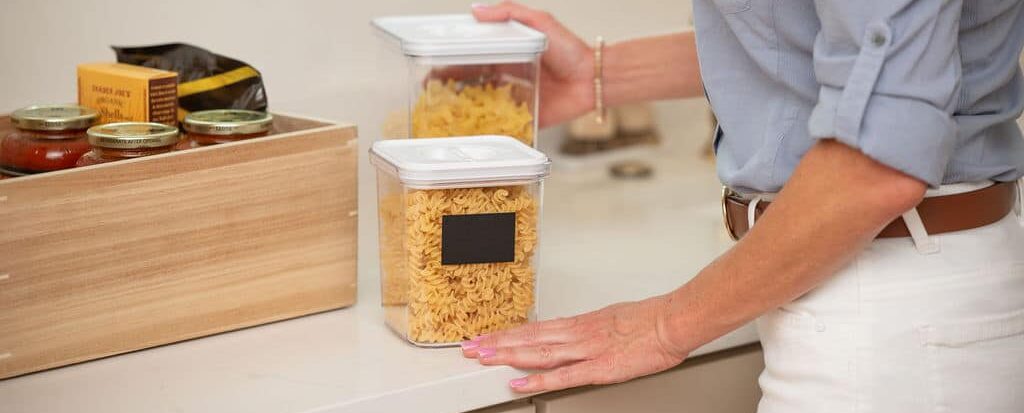 Hands organizing dry pasta into clear canisters and a bamboo storage box on a kitchen shelf.