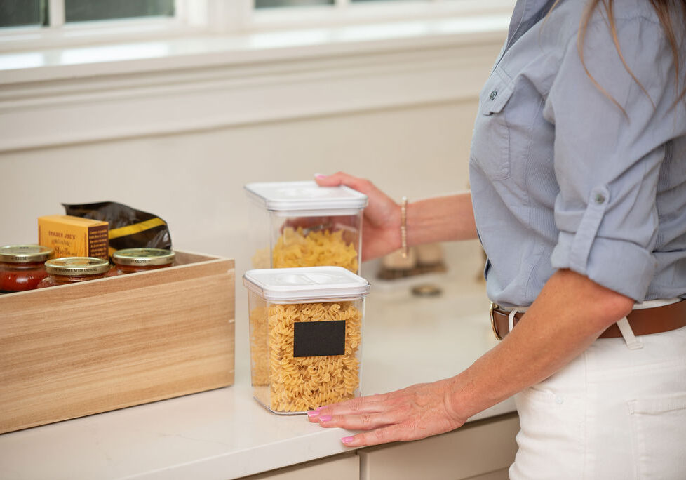 Kitchen Pantry Organization with Canisters and Bamboo Storage Hands organizing dry pasta into clear canisters and a bamboo storage box on a kitchen shelf.